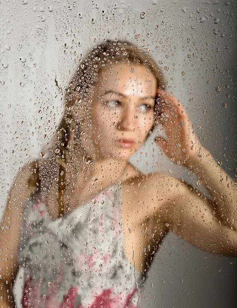 性感的年轻女子，在透明玻璃后面摆姿势，被水滴覆盖。忧郁和悲伤的女性肖像 免版税图库图片