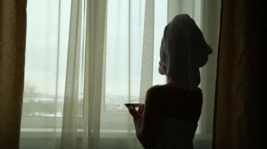 Baştan çıkarıcı kız havlu temiz vücut ile giyinmiş bir fincan sıcak çay veya kahve, ev ya da otel odasında pencere yanında ayakta tutuyor
