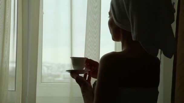 Chica seductora vestida con toalla con un cuerpo limpio está sosteniendo una taza con té o café caliente, de pie cerca de la ventana en su casa o habitación de hotel — Vídeos de Stock