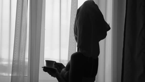 Seductive girl dressed in towel with neat body is holding a cup with hot tea or coffee, standing near the window in her home or hotel room — Αρχείο Βίντεο