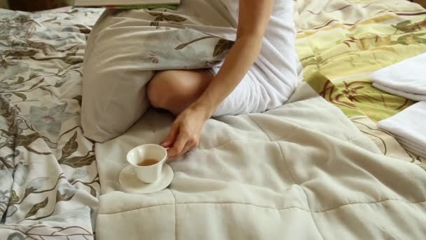 Соблазнительная девушка, одетая в полотенце с аккуратным телом, сидит на кровати, читает журнал и пьет горячий чай или кофе — стоковое видео