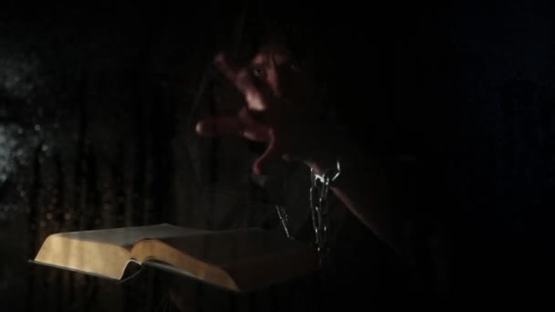 Necromancer casts spells from thick ancient book by candlelight, behind transparent glass covered by water drops on a dark background — Stockvideo