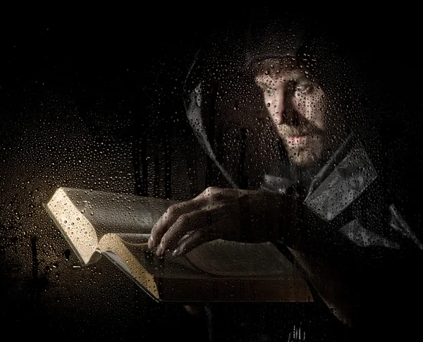 necromancer casts spells from thick ancient book, behind transparent glass covered by water drops on a dark background