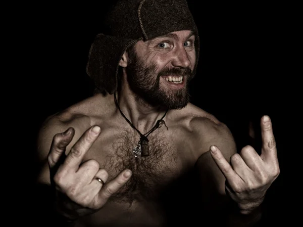 Dark portrait of scary evil sinister bearded man with smirk, shows sign of heavy metal. strange Russian man with a naked torso and a woolen hat — Stock fotografie
