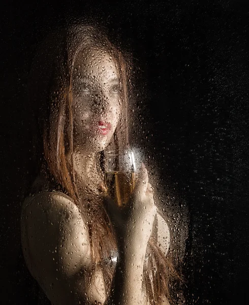 Smooth portrait of sexy model, posing behind transparent glass covered by water drops. young woman holding a glass of champagne — Stock Photo, Image