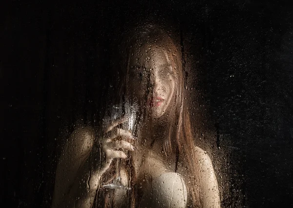 Smooth portrait of sexy model, posing behind transparent glass covered by water drops. young woman holding a glass of champagne — Stock fotografie