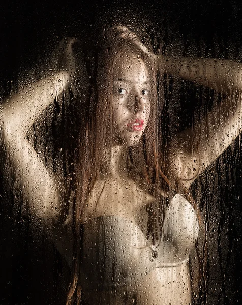 Smooth portrait of sexy model, posing behind transparent glass covered by water drops. — Stock Photo, Image