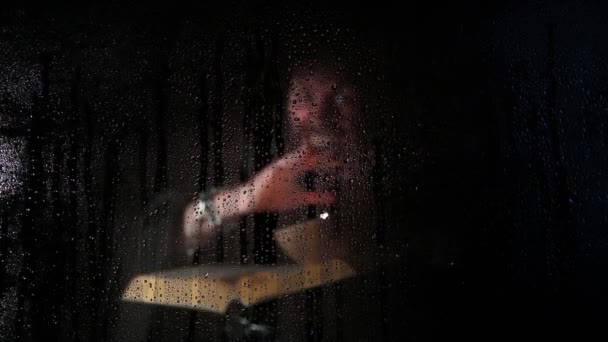 Некромант накладывает заклинания из толстой древней книги при свечах, за прозрачным стеклом, покрытым каплями воды на темном фоне — стоковое видео