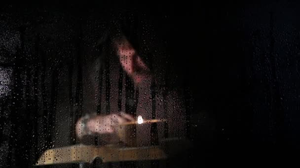 Некромант накладывает заклинания из толстой древней книги при свечах, за прозрачным стеклом, покрытым каплями воды на темном фоне — стоковое видео