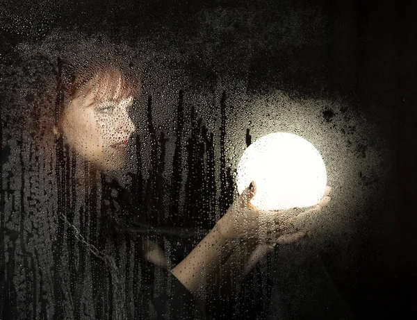 Retrato liso de modelo femenino, posando detrás de vidrio transparente cubierto por gotas de agua. mujer sosteniendo gran bola brillante —  Fotos de Stock