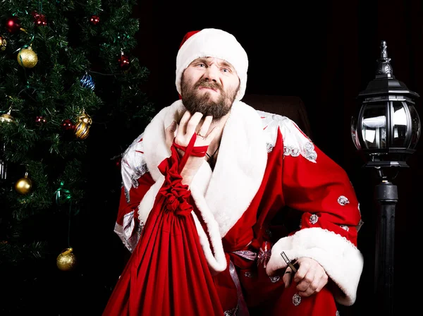 Cattivo brutale Babbo Natale tenendo la borsa con regali e graffiando la barba sullo sfondo dell'albero di Natale — Foto Stock