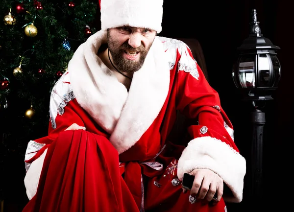 Mal brutal Santa Claus sosteniendo la bolsa con regalos en el fondo del árbol de Navidad — Foto de Stock