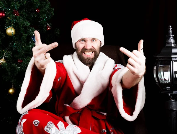 Bad brutal Santa Claus smiles and showing middle finger sign on the background of Christmas tree — Stock Photo, Image
