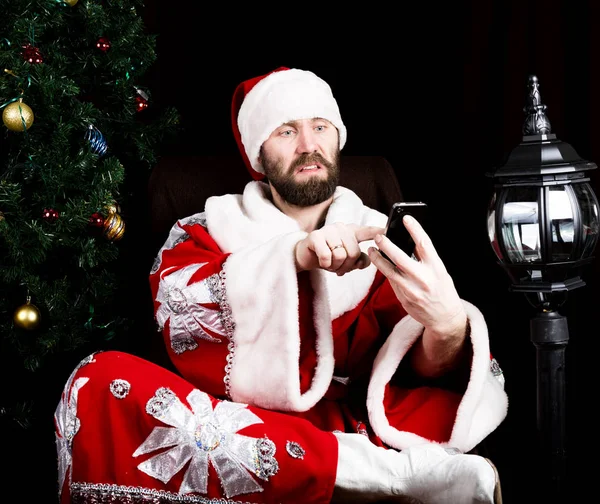 Bad brutal Santa Claus holding the bag with gifts and dissatisfied talking phone on the background of Christmas tree — Stock Photo, Image