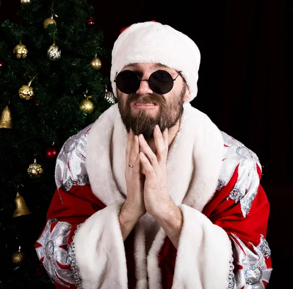 Bad rastoman Santa Claus holding the bag with gifts and scratching his beard on the background of Christmas tree — Stock Photo, Image