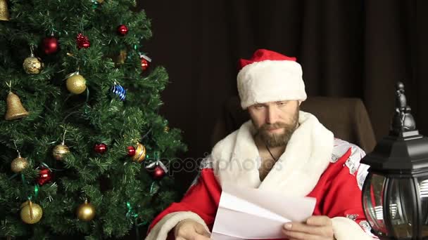 Ruim brutal Papai Noel descontente lê carta com os desejos, no fundo da árvore de Natal — Vídeo de Stock