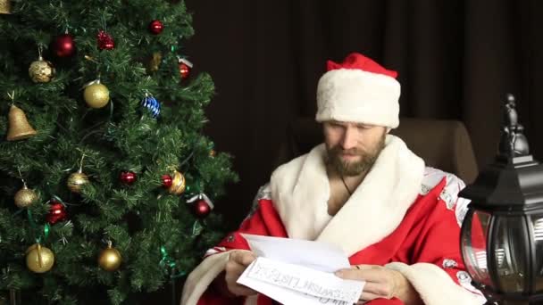 Bad brutal Santa Claus discontentedly reads letter with the wishes, on the background of Christmas tree — Stock Video