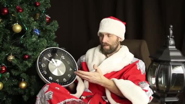 Mal brutal Santa Claus sonriendo y muestra el reloj, de cinco minutos a doce, en el fondo del árbol de Navidad — Vídeos de Stock