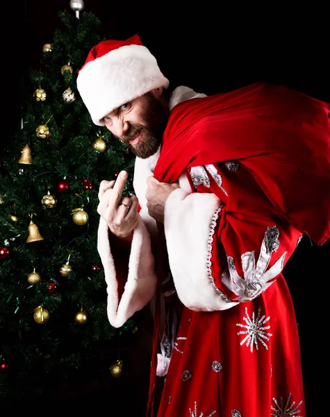 Bad brutal Santa Claus carries a bag, showing middle finger and smiling spitefully, on the background of Christmas tree — Stock Photo, Image