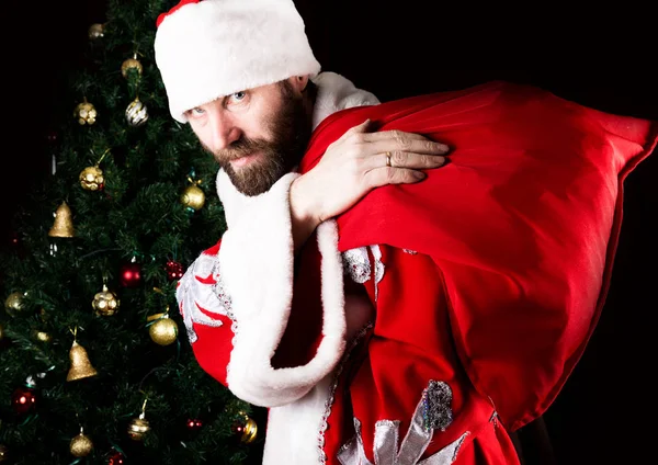 Bad brutal Santa Claus carries a bag and smiling spitefully, on the background of Christmas tree — Stock Photo, Image