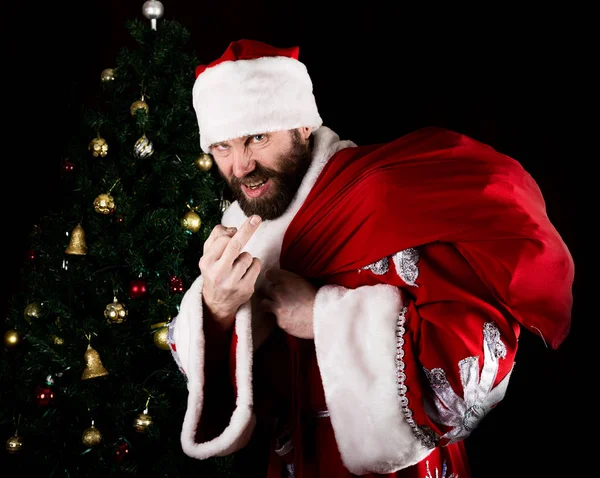 中指を示し、クリスマス ツリーの背景に、意地悪く笑みを浮かべて悪い残忍なサンタ クロースが袋を運ぶ — ストック写真