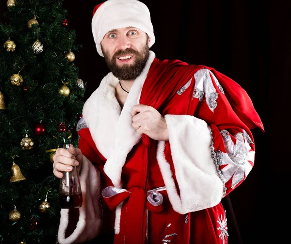 Bad brutal Santa Claus carries a bag, smiling spitefully and drinking brandy from a bottle, on the background of Christmas tree — Stock Photo, Image