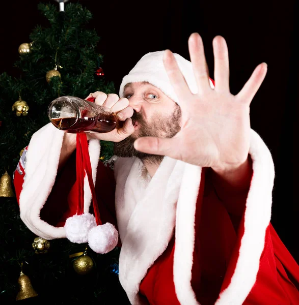 Huono brutaali joulupukki hymyilee innokkaasti, juo brandyä pullosta ja ojennetusta käsivarresta joulukuusen taustalla. — kuvapankkivalokuva