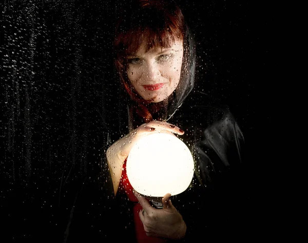 Портрет жіночої моделі, що позує за прозорим склом, покритим краплями води. жінка тримає великий сяючий м'яч — стокове фото