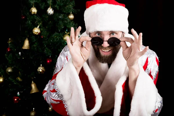 Cattivo rastoman Babbo Natale tiene bicchieri rotondi e sorrisi, sullo sfondo dell'albero di Natale. diverse emozioni divertenti — Foto Stock