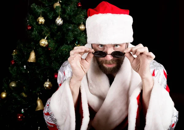 Cattivo rastoman Babbo Natale tiene bicchieri rotondi e sorrisi, sullo sfondo dell'albero di Natale. diverse emozioni divertenti — Foto Stock