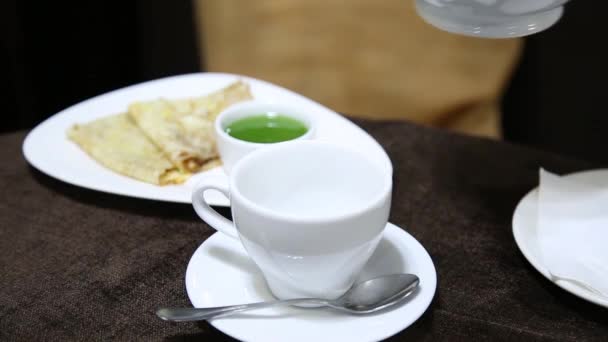 Nalévání čaje do bílý šálek na stole z porcelánové konvice. želé a palačinka na pozadí — Stock video