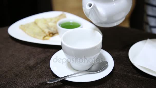 Выливание чая в белый стакан на стол из фарфорового чайника. желе и блины на заднем плане — стоковое видео