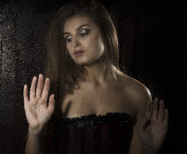 Портрет чувственной молодой сексуальной женщины, носящей корсет, трогающей волосы и позирующей за прозрачным стеклом, покрытым каплями воды . — стоковое фото
