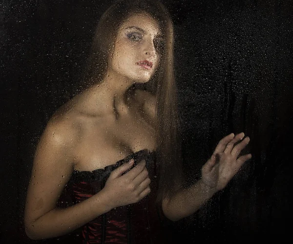 Portrait of a sensual young sexy woman wearing corset, touching her hair and posing behind transparent glass covered by water drops. — Stock Photo, Image