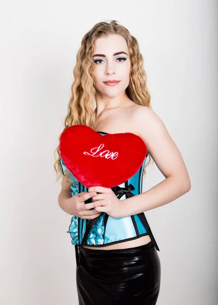 Young and beautiful curly girl in blue corset holding a red heart pillow — Stock Photo, Image