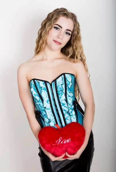 Young and beautiful curly girl in blue corset holding a red heart pillow — Stock Photo, Image