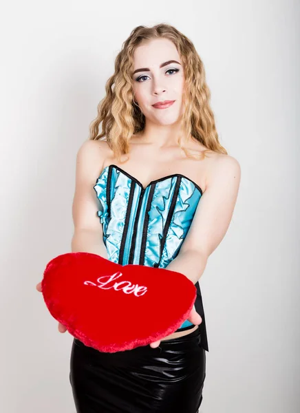 Mladá a krásná kudrnatá dívka v modrém korzet, držící červené srdce polštář — Stock fotografie