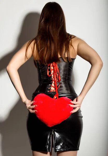 Mladá krásná dívka v Kožený korzet se šněrováním a sukni, držící červené srdce polštář — Stock fotografie