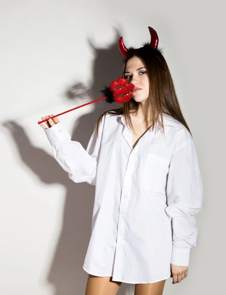 Joven chica en un mans blanco camisa con cuernos rojos sosteniendo tridente y se ve como bonito diablo — Foto de Stock