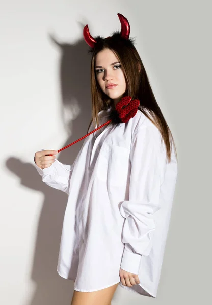 Joven chica en un mans blanco camisa con cuernos rojos sosteniendo tridente y se ve como bonito diablo — Foto de Stock