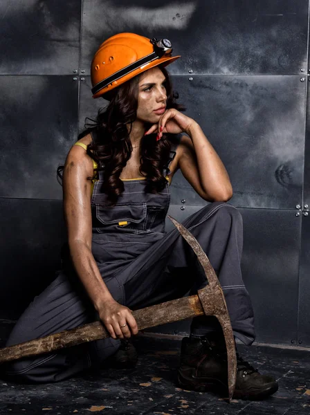 Sexy žena horník dělník s krumpáčem v kombinéze jeho nahé tělo, sedící na podlaze na pozadí ocelová zeď — Stock fotografie