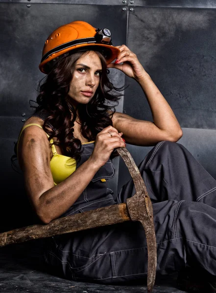 Sexy žena horník dělník s krumpáčem v kombinéze jeho nahé tělo, sedící na podlaze na pozadí ocelová zeď — Stock fotografie