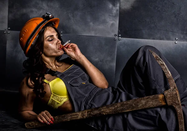 Sexy operaia minatore con piccone, in tuta sopra il suo corpo nudo, seduto sul pavimento sullo sfondo di parete in acciaio e fumo — Foto Stock