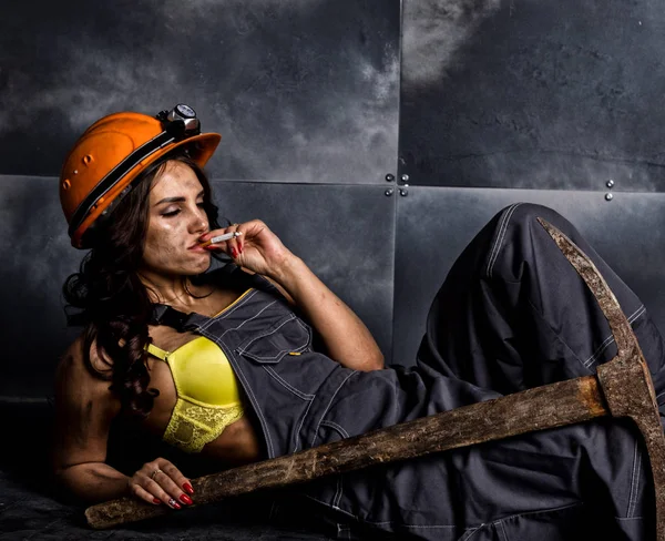 sexy female miner worker with pickaxe, in coveralls over his naked body, sitting on the floor on backdrop of steel wall and smoking