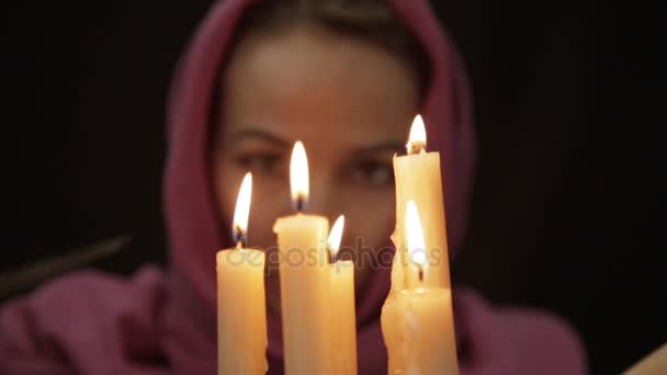 Una mujer con un pañuelo en la cabeza haciendo el ritual mágico. primer plano varias velas y libro viejo. halloween o concepto de religión — Vídeo de stock