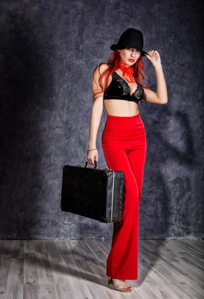 Bella donna rossa in pantaloni rossi, reggiseno e mans cappello in piedi e in possesso di vecchia valigia — Foto Stock