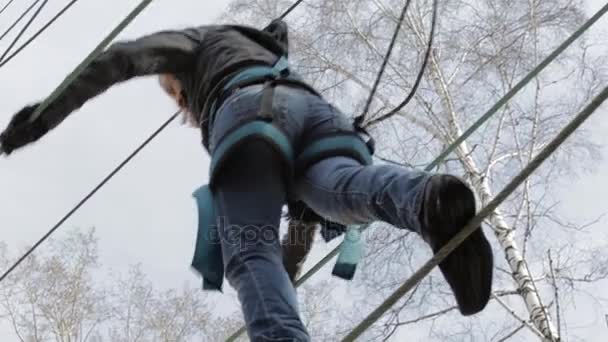 Jovem alpinista caminha pela ponte de corda pingente no curso de cordas altas no parque extremo. Conceito de escalada — Vídeo de Stock