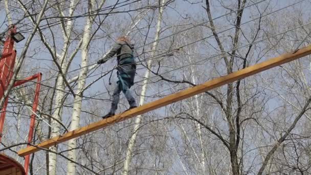 Молодая альпинистка прогуливается по бревенчатому мосту на высоких канатах в экстремальном парке — стоковое видео