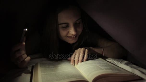 Молодая девушка использует фонарик в темноте для чтения книги под одеялом — стоковое видео