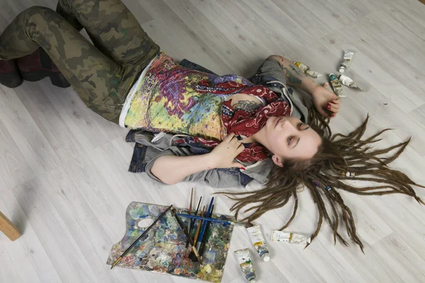 Atraktivní mladá zpěvačka s dredy leží na podlaze, drží štětce, jsou trubky s barvami v okolí — Stock fotografie